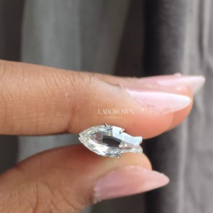 May include: A clear, pear-shaped gemstone with a brilliant cut, held between fingers. The gemstone is set in a silver or white metal. The text "LABGROWN GERMANY" is visible in the image.