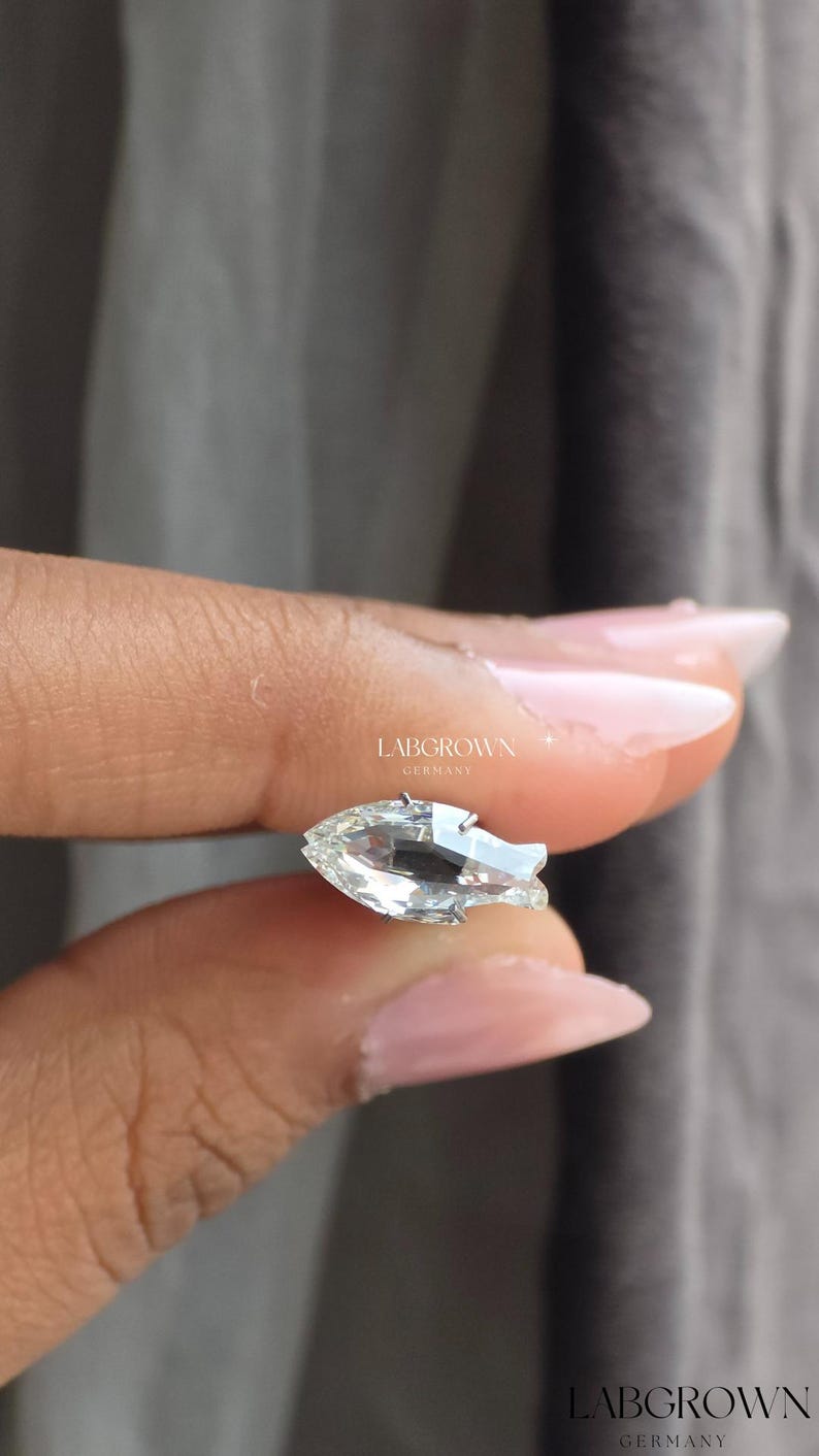 May include: A clear, pear-shaped gemstone with a brilliant cut, held between fingers. The gemstone is set in a silver or white metal. The text "LABGROWN GERMANY" is visible in the image.