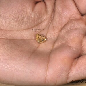 May include: A yellow heart-shaped gemstone, lab-grown in Germany, is displayed on a hand.