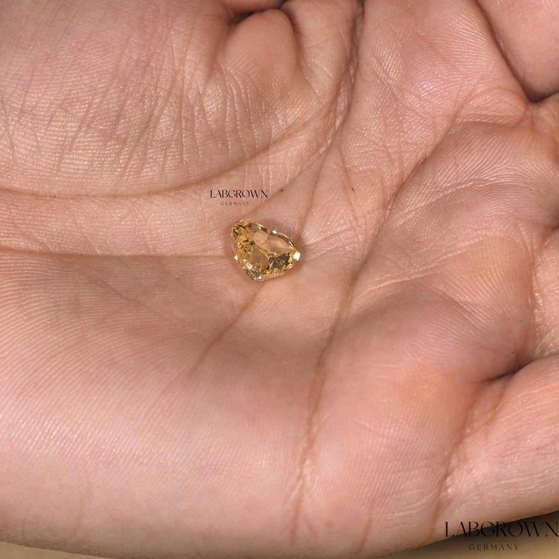 May include: A yellow heart-shaped gemstone, lab-grown in Germany, is displayed on a hand.