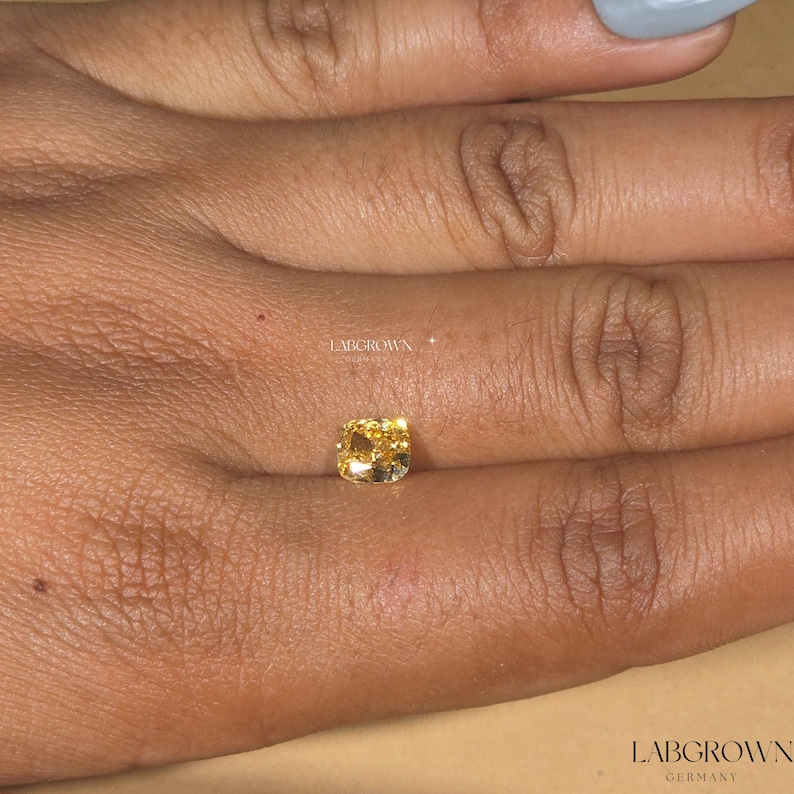 May include: A cushion-cut yellow lab-grown diamond, set in a ring, on a person's finger. LABGROWN GERMANY is visible in the image.