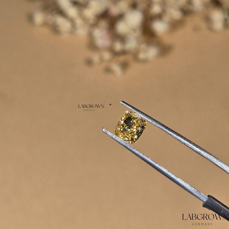 May include: A cushion-cut, yellow lab-grown diamond held by tweezers. The diamond is a light yellow color with a slight green hue. LABGROWN GERMANY is printed on the tweezers.