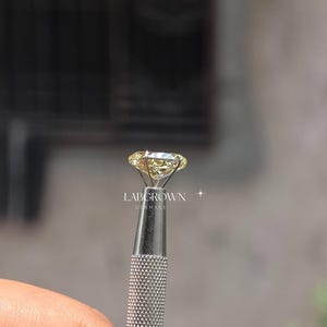 以下が含まれることがあります： 銀色のツールで保持された、大きく丸い黄色のダイヤモンド。 ダイヤモンドはカットされており、光を反射しています。 ツールは手で持たれています。 テキスト「LABGROWN GERMANY」が表示されています。