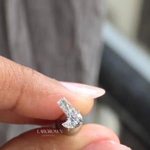 Puede incluir: Primer plano de un pendiente de diamante cultivado en laboratorio. El pendiente presenta un diamante transparente de corte rectangular engastado en metal plateado. El texto "LABGROWN GERMANY" es visible.