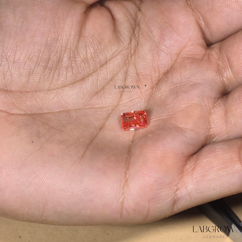 Puede incluir: Una peque&ntilde;a gema naranja rectangular con talla brillante se muestra en la palma de una mano. La gema es de un tono naranja vibrante, y las facetas captan la luz. El texto "LABGROWN GERMANY" es visible en la imagen.