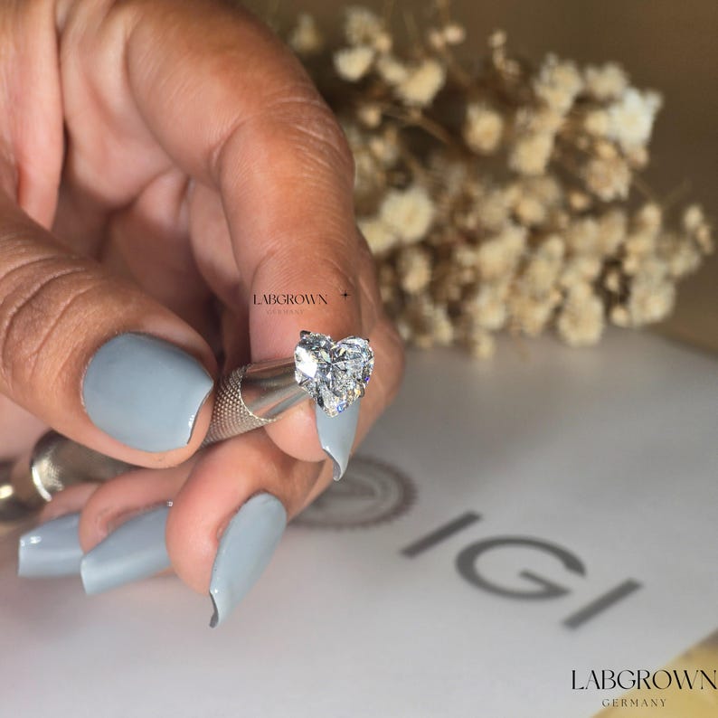 May include: A heart-shaped lab-grown diamond held in a tweezers. The diamond is clear and sparkly. The background is a white surface with the text "IGI LABGROWN GERMANY" printed on it.