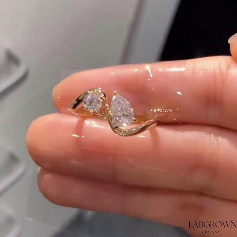 May include: A gold ring with two pear-shaped diamonds set in a twisted band. The ring is on a finger and the brand name "LABGROWN GERMANY" is visible.