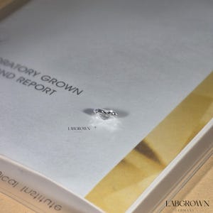 May include: A clear, lab-grown diamond in a heart shape, resting on a white surface with the text "Laboratory Grown and Report" and "Labgrown" printed on it.