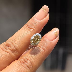 以下が含まれることがあります： シャンパンカラーの中心とシルバーカラーのセッティングが施された、オーバルカットのラボグロウンダイヤモンド。指でダイヤモンドを持っています。「LABGROWN GERMANY」の文字が見えます。