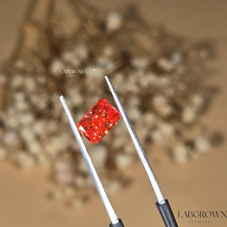 Puede incluir: Una radiante gema roja, de corte rectangular, cultivada en laboratorio, es sostenida por unas pinzas. La gema brilla con facetas internas, reflejando la luz. La imagen incluye el texto "LABGROWN GERMANY".