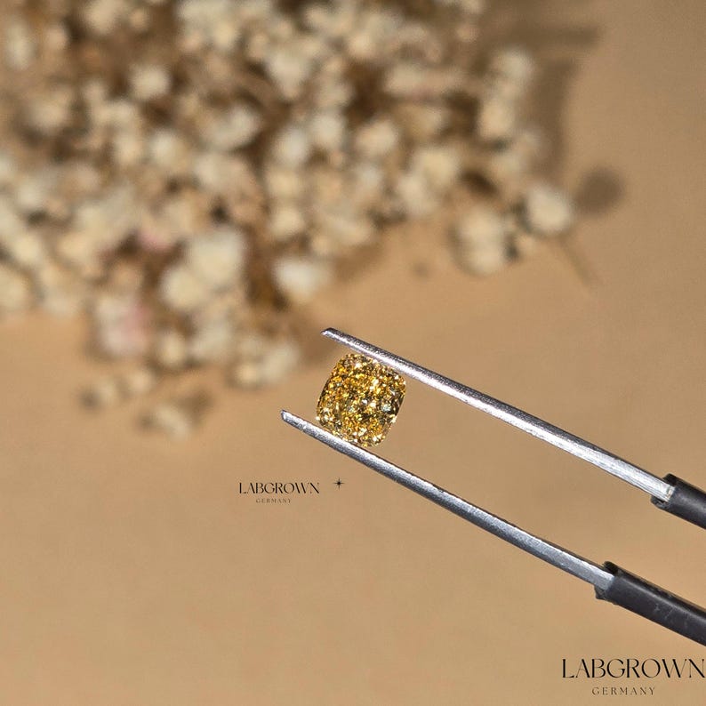 May include: A yellow lab-grown diamond held in tweezers. The diamond is a cushion cut and has a bright, golden color. The diamond is set against a background of dried flowers.