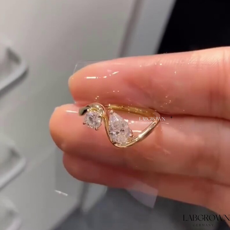 May include: A gold ring with two pear-shaped diamonds set in a curved band. The ring is on a finger and the text "LABGROWN GERMANY" is visible in the background.