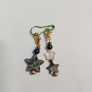 Op de afbeelding: Twee gouden stervormige steekmarkeerders met zwarte kralen en een groene veiligheidsspeld. De sterren zijn blauw en wit met gouden accenten.