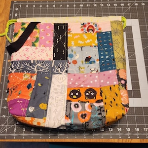 May include: A colorful patchwork drawstring bag with a black strap. The bag is made of various fabrics with different patterns, including cats, stars, and trees. The bag is approximately 12 inches wide and 14 inches tall.