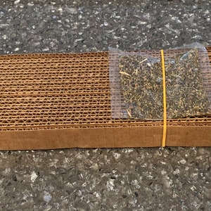 Può includere: Un mucchio di fogli di cartone marrone tenuti insieme da un elastico giallo. Un piccolo sacchetto di plastica trasparente pieno di erbe secche è posto sopra il cartone.