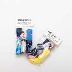 Può includere: Due sacchetti di plastica trasparenti pieni di giocattoli annodati con erba gatta colorati. I sacchetti hanno etichette che recitano "Catnip Knots Catnip Filled Toys!" e includono l'indirizzo del sito web "www.CatScratcherGuy.com".