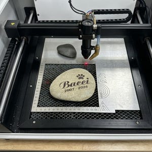 K&ouml;nnte beinhalten: Eine Lasergravurmaschine mit schwarzem Metallrahmen und einem Laserkopf &uuml;ber einem Wabenbett. Ein heller Stein mit der Gravur Bacci 2007-2026 und einem Pfotenabdruck liegt auf dem Gitter, daneben befindet sich ein schlichter, dunkelgrauer Stein.