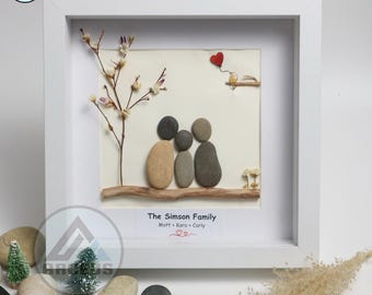 May include: A framed pebble art piece depicting a family of three with a small tree and a bird with a red heart balloon. The text below reads "The Simson Family Matt + Kara + Carly".