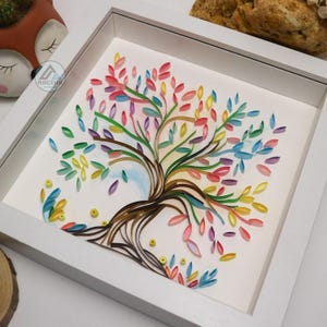May include: A framed paper art piece depicting a colorful tree with a brown trunk and branches. The leaves are made of curled paper in shades of pink, blue, green, yellow, and purple.