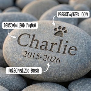 May include: A grey engraved stone featuring the name "Charlie", the years "2015-2026", and a paw print icon. The stone is set amongst other grey stones, giving a natural, outdoor feel.