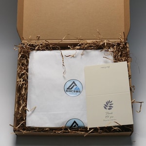 May include: A brown cardboard box with a white tissue paper lining. The box contains a thank you card with a leaf design and the text "Thank you. May you have a lovely time." The box also has a sticker with a blue and white logo that says "Arceus Handmade".