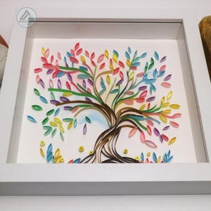 May include: A framed paper art piece depicting a colorful tree with brown branches and leaves in shades of pink, blue, green, yellow, and purple. The tree is set against a white background with a light blue watercolor wash.