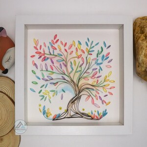 May include: A framed artwork featuring a colorful paper quilled tree with a rainbow of colors in the leaves. The tree is set against a white background and is framed in a white frame.