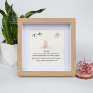 May include: A framed piece of art with a white background and a message about a daughter. The message is written in a cursive font and includes the name "Emily". The art is decorated with small, colorful stones and a white flower.
