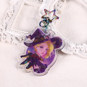Può includere: Un portachiavi con un personaggio anime di colore viola e nero con un cappello da strega e un ciondolo a forma di stella. Il personaggio ha i capelli biondi e gli occhi azzurri.