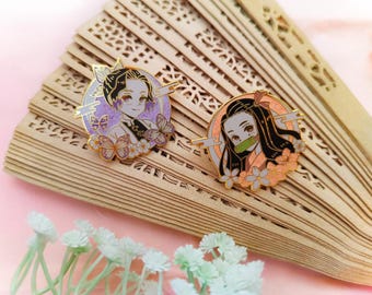May include: Two enamel pins featuring anime characters with floral details. The first pin features a female character with purple hair and a butterfly design. The second pin features a female character with black hair and a pink flower design.