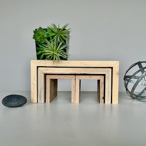 Może przedstawiać: Trzy drewniane stoliki wkładane, każdy z prostokątnym blatem i otwartymi bokami, ułożone w kolejności malejącej. Stoliki są wykonane z naturalnego drewna o jasnobrązowym wykończeniu. Mała roślina w doniczce stoi na największym stoliku.