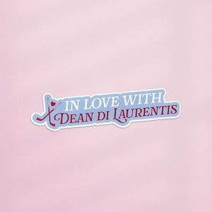 May include: A sticker with the text "IN LOVE WITH DEAN DI LAURENTIS" in a stylized font. The text is in burgundy and light blue, with a hockey stick and heart graphic. The sticker is on a pink background.