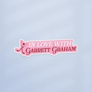 May include: A pink sticker with the text "IN LOVE WITH Garrett Graham" in white and red lettering. The sticker features two crossed hockey sticks and a heart. The sticker has a white border and is on a light blue background.