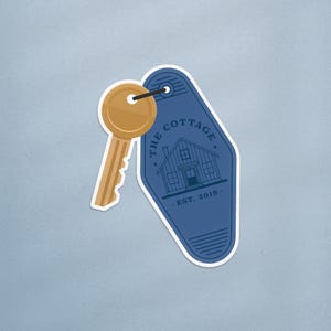 Peut inclure: Une clé dorée attachée à un porte-clés bleu. Le porte-clés porte l'inscription "THE COTTAGE", une illustration de maison et le texte "EST. 2019."