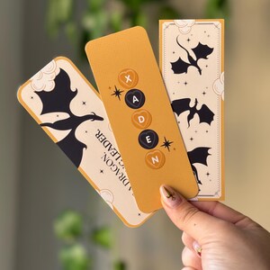 May include: Three yellow bookmarks with black dragon silhouettes and a white background. The middle bookmark has the letters "XADEN" in black circles with a heart shape around each letter.