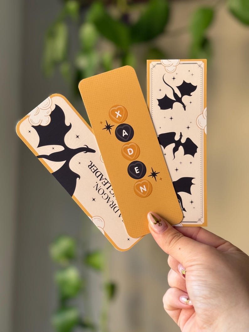 May include: Three yellow bookmarks with black dragon silhouettes and a white background. The middle bookmark has the letters "XADEN" in black circles with a heart shape around each letter.