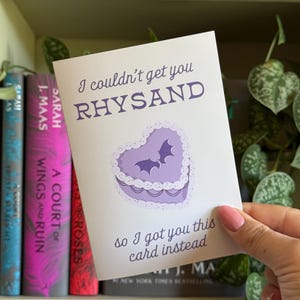 May include: A greeting card with the text "I couldn't get you RHYSAND, so I got you this card instead." The card features a purple heart-shaped cake with two bat silhouettes. Books are visible in the background.