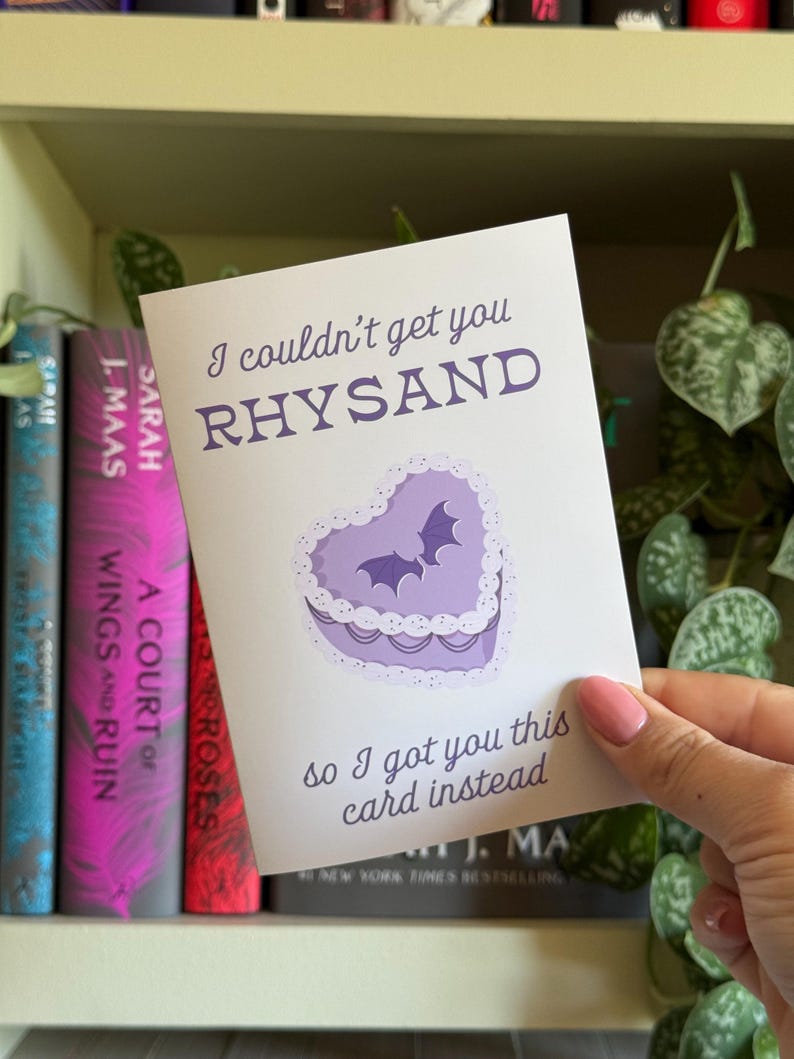 May include: A greeting card with the text "I couldn't get you RHYSAND, so I got you this card instead." The card features a purple heart-shaped cake with two bat silhouettes. Books are visible in the background.