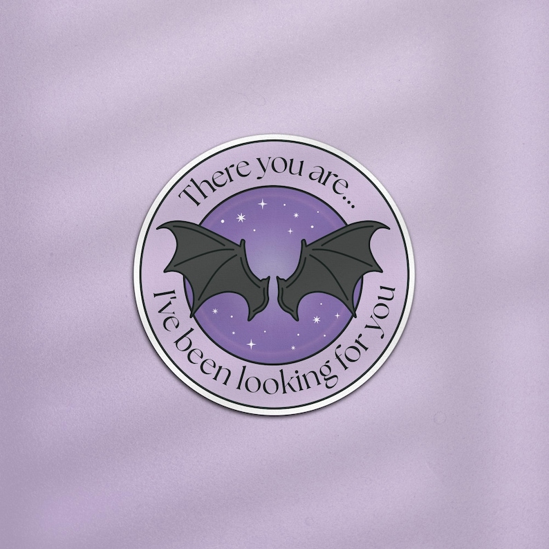 May include: A round sticker with a purple background and a silver border. The sticker features a pair of black bat wings in the center, with the text "There you are... I've been looking for you" in a circular pattern around the wings.