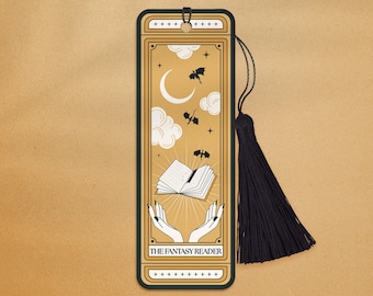 Può includere: Un segnalibro nero con sfondo dorato e un disegno di un libro con le ali che vola nel cielo con una luna crescente e stelle. Il segnalibro ha un fiocco nero e il testo "THE FANTASY READER".
