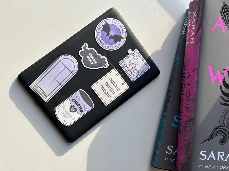 Pode incluir: Um leitor de e-books preto com v&aacute;rios adesivos nele. Os adesivos s&atilde;o roxos e brancos e apresentam um morcego, uma janela, uma lata de sopa, um cora&ccedil;&atilde;o e um selo. O leitor de e-books est&aacute; sobre uma superf&iacute;cie branca.