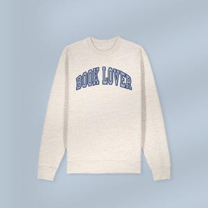 May include: A cream-coloured sweatshirt featuring the text "BOOK LOVER" in blue, arched across the chest. The sweatshirt has a crew neck and long sleeves. The background is a light blue.