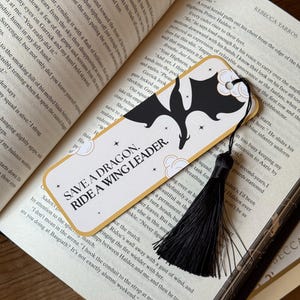 May include: A gold and black bookmark with a black tassel. The bookmark features a black silhouette of a dragon with a white cloud and stars. The text on the bookmark reads "Save a Dragon. Ride a Wing Leader."