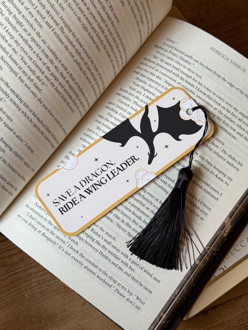May include: A gold and black bookmark with a black tassel. The bookmark features a black silhouette of a dragon with a white cloud and stars. The text on the bookmark reads "Save a Dragon. Ride a Wing Leader."
