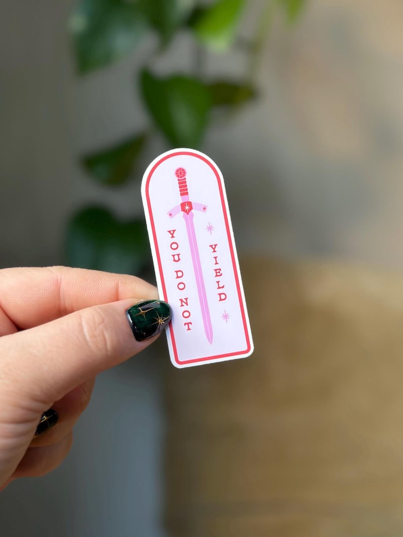 May include: A sticker with a pink sword design and the words "YOU DO NOT YIELD" in pink text. The sticker has a white background with a red border and is held by a hand with a dark green nail polish.