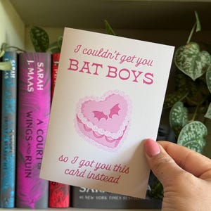 May include: A greeting card with the text "I couldn't get you BAT BOYS so I got you this card instead." The card features a pink heart-shaped cake with bats. The card is held in front of a bookshelf with books.