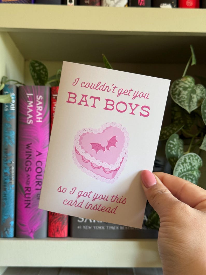 May include: A greeting card with the text "I couldn't get you BAT BOYS so I got you this card instead." The card features a pink heart-shaped cake with bats. The card is held in front of a bookshelf with books.