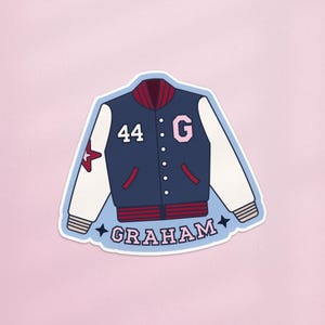May include: A sticker depicting a navy blue varsity jacket with white sleeves. The jacket features a red collar and pocket details, with the number 44 and the letter G in pink. The name "GRAHAM" is written in pink script below.