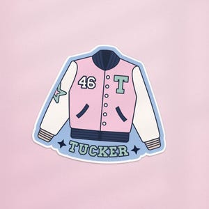 Puede incluir: Una pegatina de chaqueta universitaria rosa y blanca con el número 46 y la letra T. La chaqueta tiene cuello y bolsillos azules, con el nombre "Tucker" impreso en la parte inferior. Una estrella verde está en la manga izquierda.