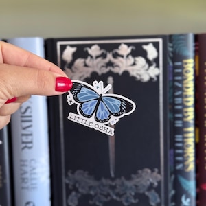 K&ouml;nnte beinhalten: Ein Aufkleber mit einem blau-schwarzen Schmetterlingsdesign und dem Text "LITTLE OSHA". Der Aufkleber wird vor einem dunkelblauen Buch mit silbernen floralen Akzenten und anderen B&uuml;chern auf einem Regal gehalten.